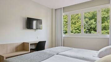 A hotel room with two beds, a TV mounted on the wall, a desk, and a window with a view of trees.