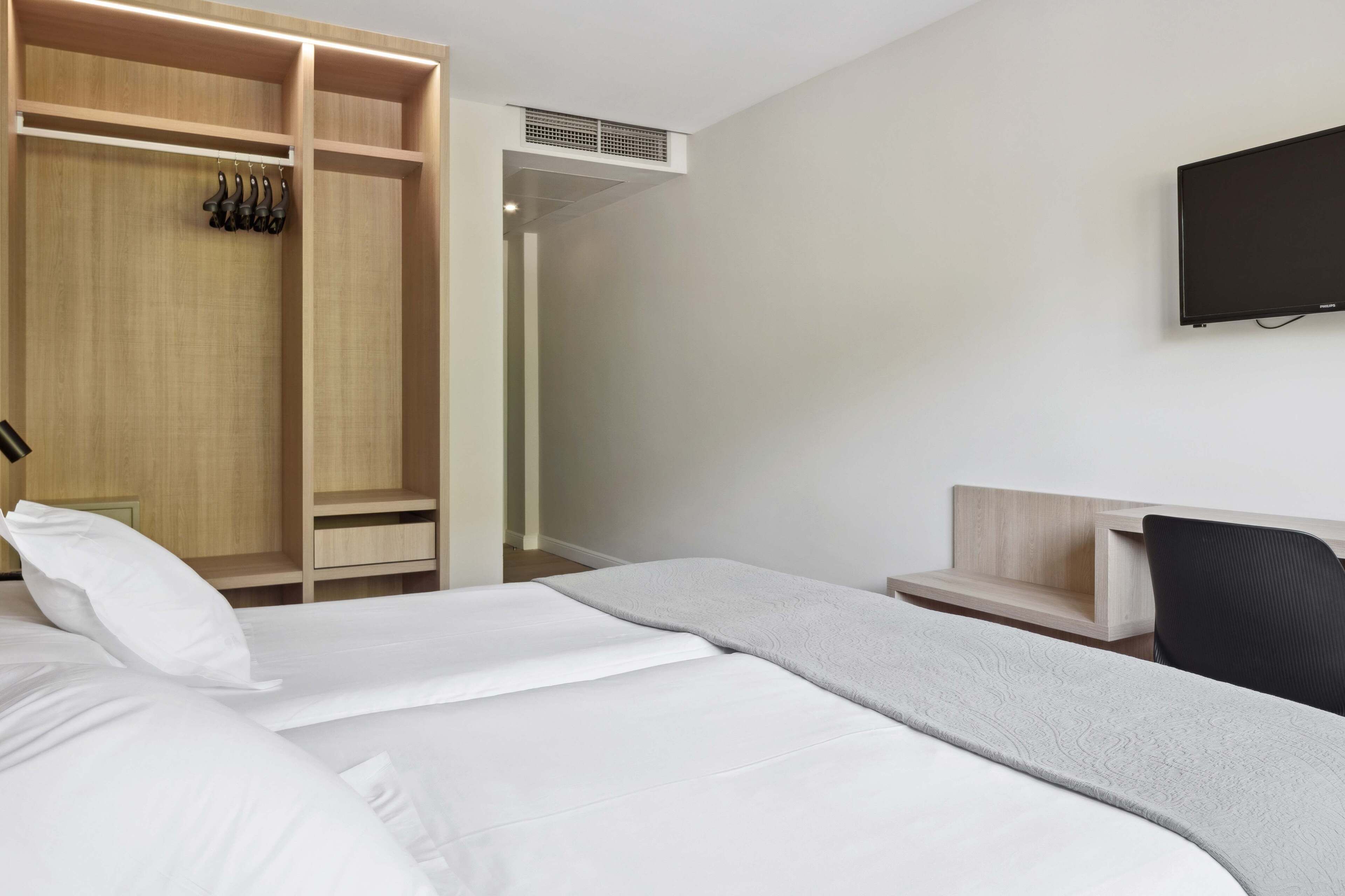 A hotel room with a bed, a wooden wardrobe, a desk, a chair, and a TV mounted on the wall.