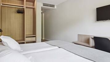 A hotel room with a bed, a wooden wardrobe, a desk, a chair, and a TV mounted on the wall.