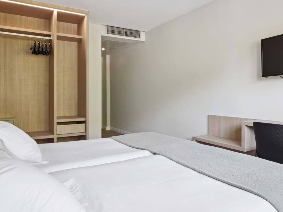 A hotel room with a bed, a wooden wardrobe, a desk, a chair, and a TV mounted on the wall.