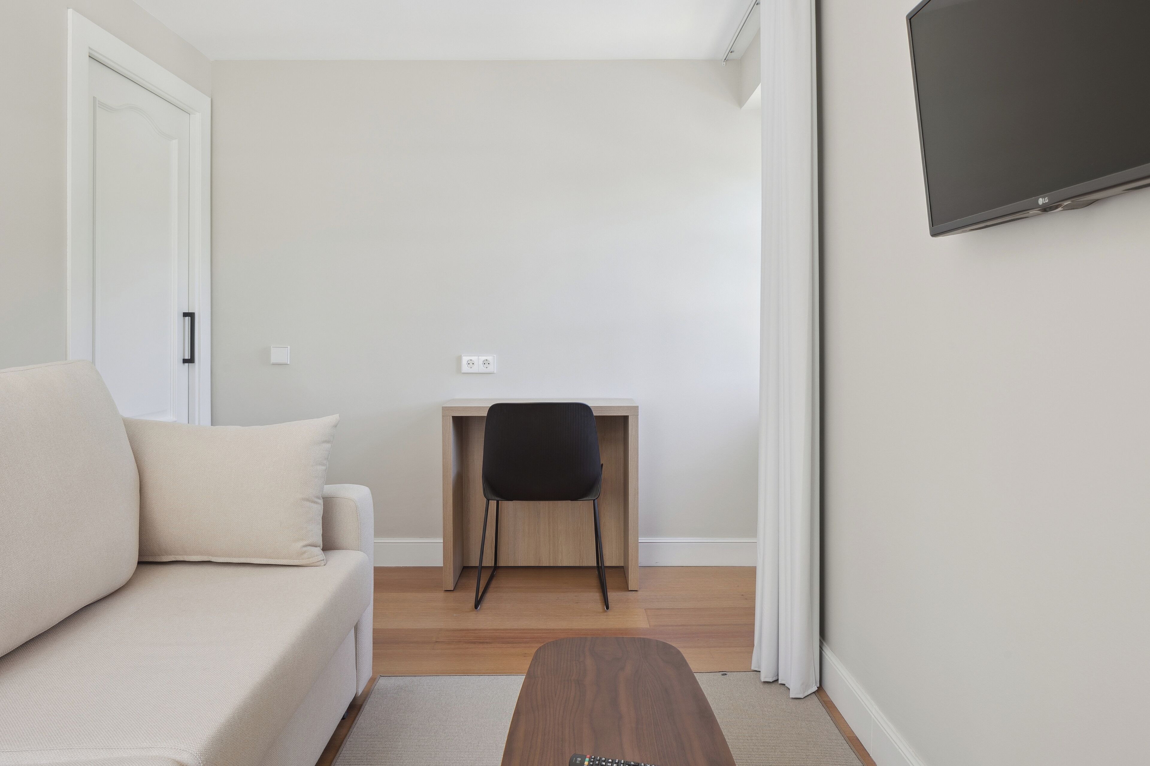 A modern living room with a sofa, a wooden desk, a black chair, and a flat-screen TV mounted on the wall.