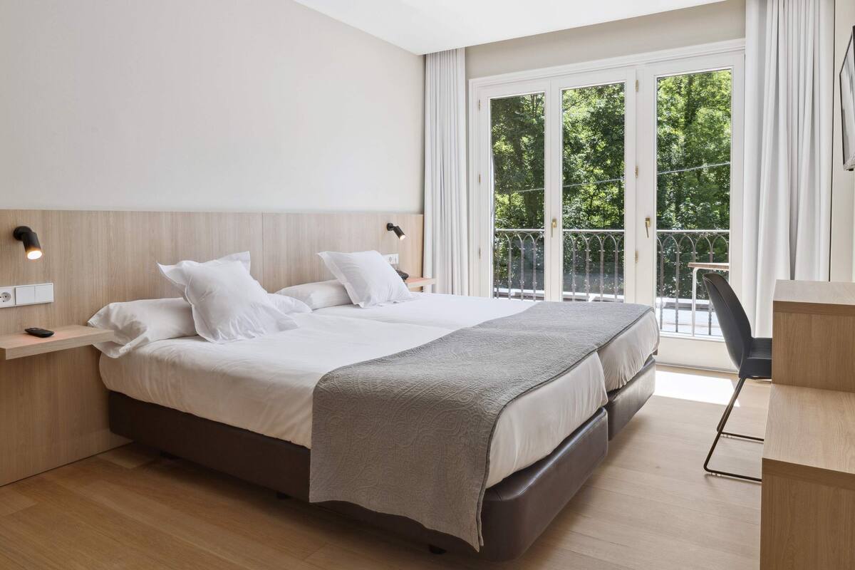 Habitación de hotel moderna con una cama grande, un escritorio y un balcón con vistas a zonas verdes.