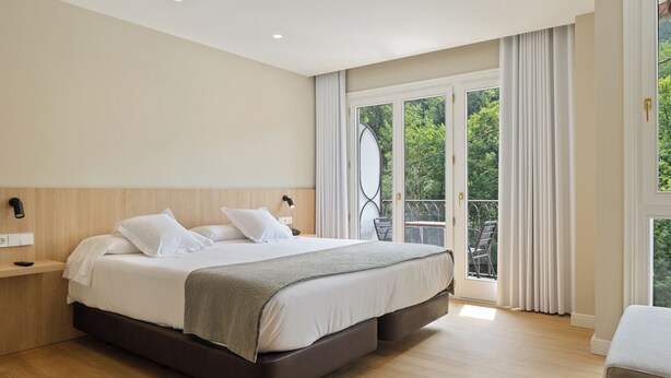 A modern bedroom with a large bed, a bedside table, and a view of greenery through a sliding glass door. - Hotel Venta Etxalar (Etxalar)