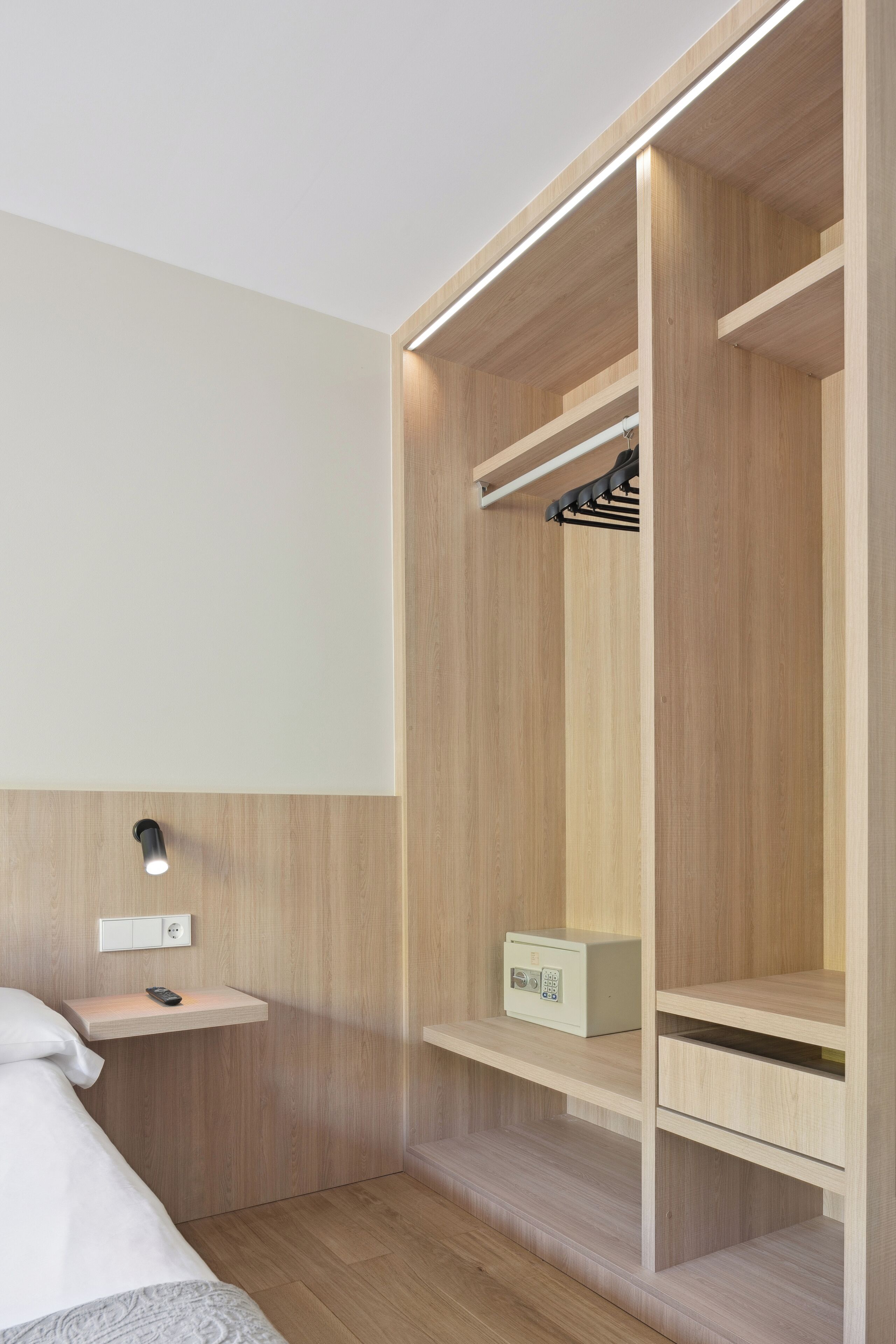 Una habitación de hotel moderna con un armario de madera, una mesita de noche con un control remoto y una pequeña caja en la estantería.