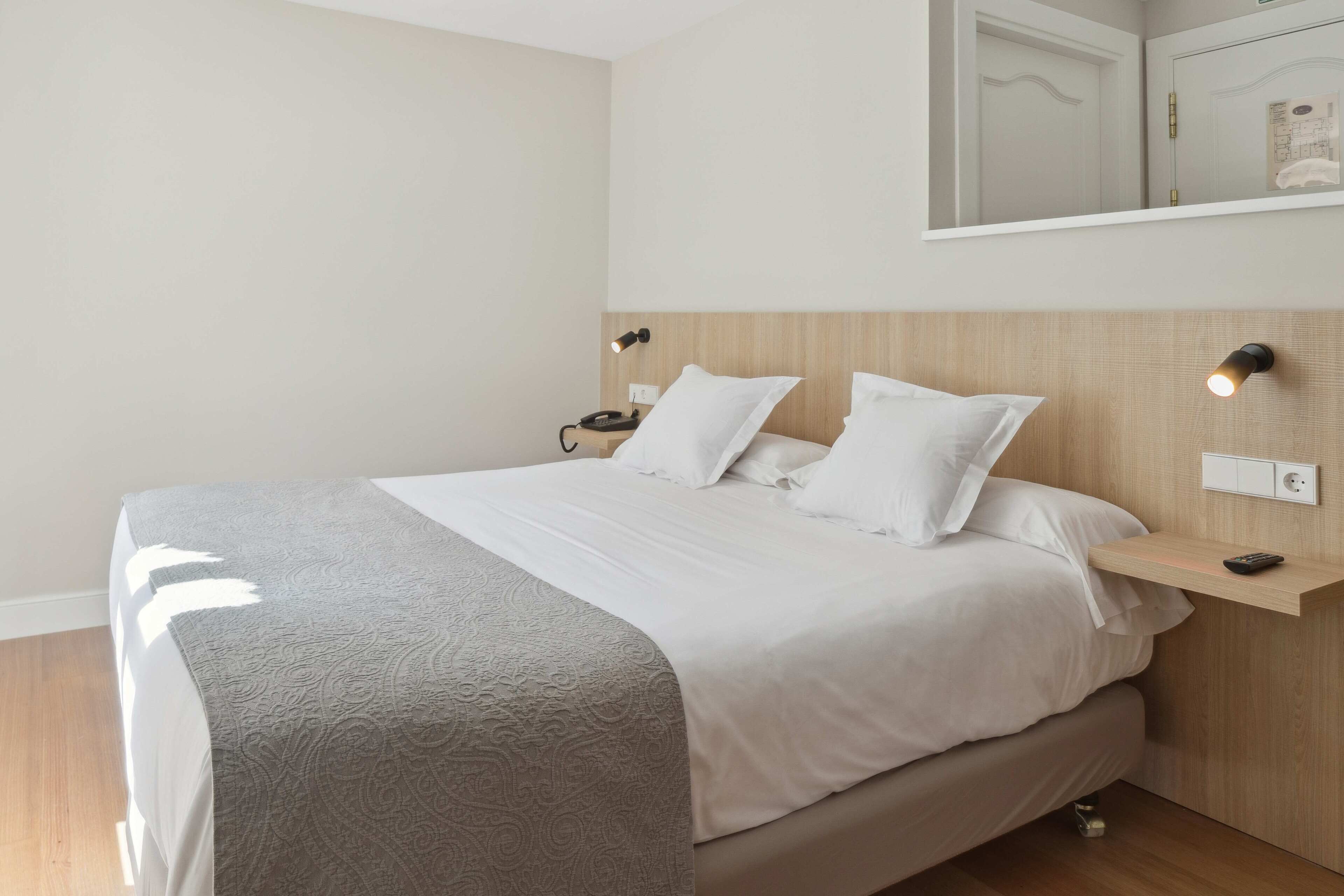 A neatly made bed with a grey patterned bedspread, a wooden headboard, bedside tables with lamps, and a wall-mounted mirror.
