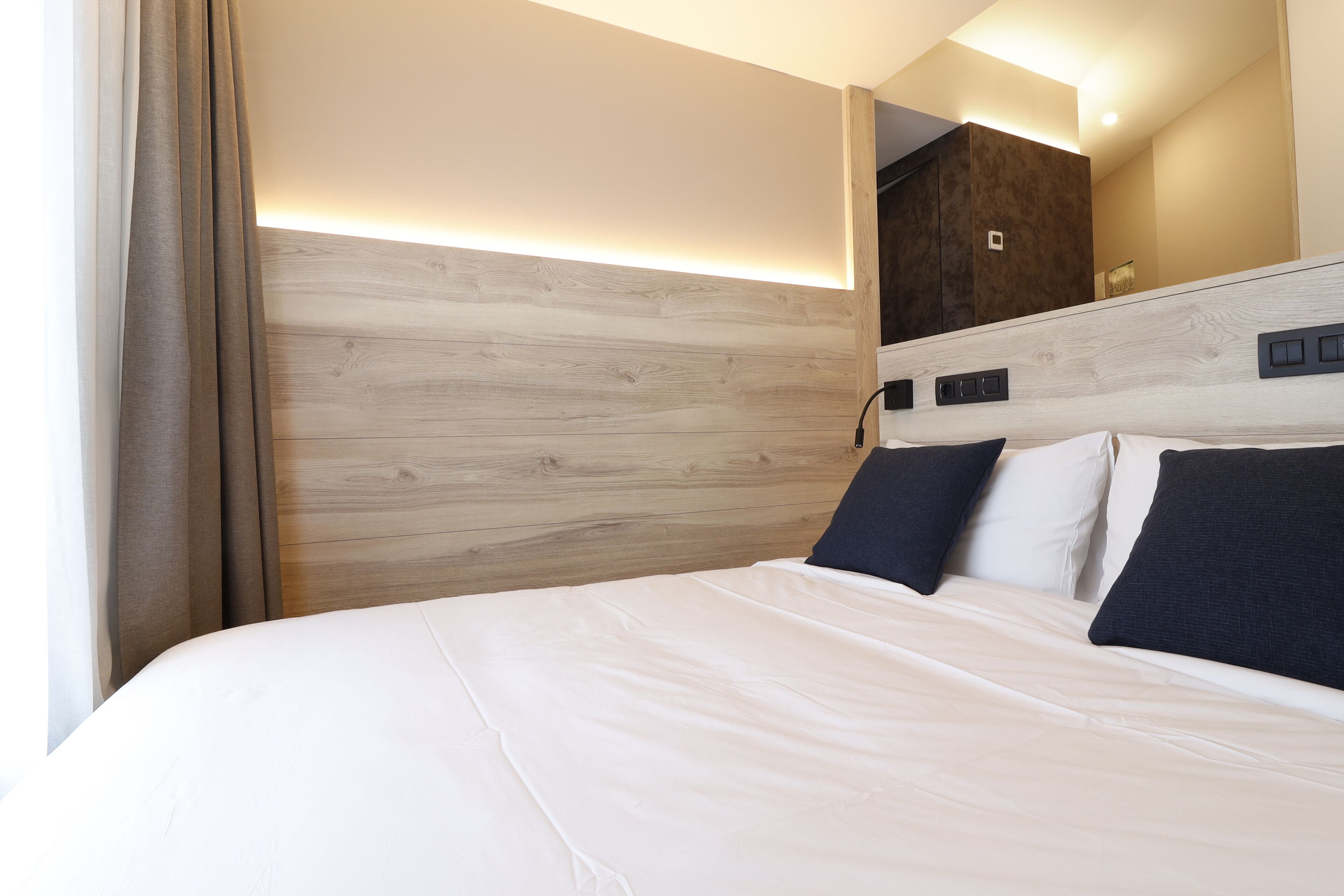 A modern hotel room with a wooden headboard, a large bed with white bedding, and a built-in wardrobe with a mirror.