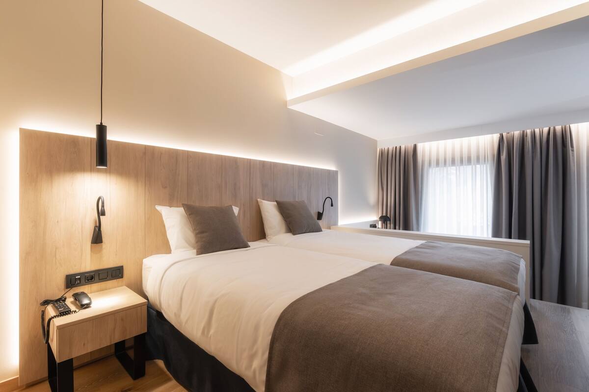 A modern hotel room with a large bed, bedside tables, a desk, and a large window with curtains.