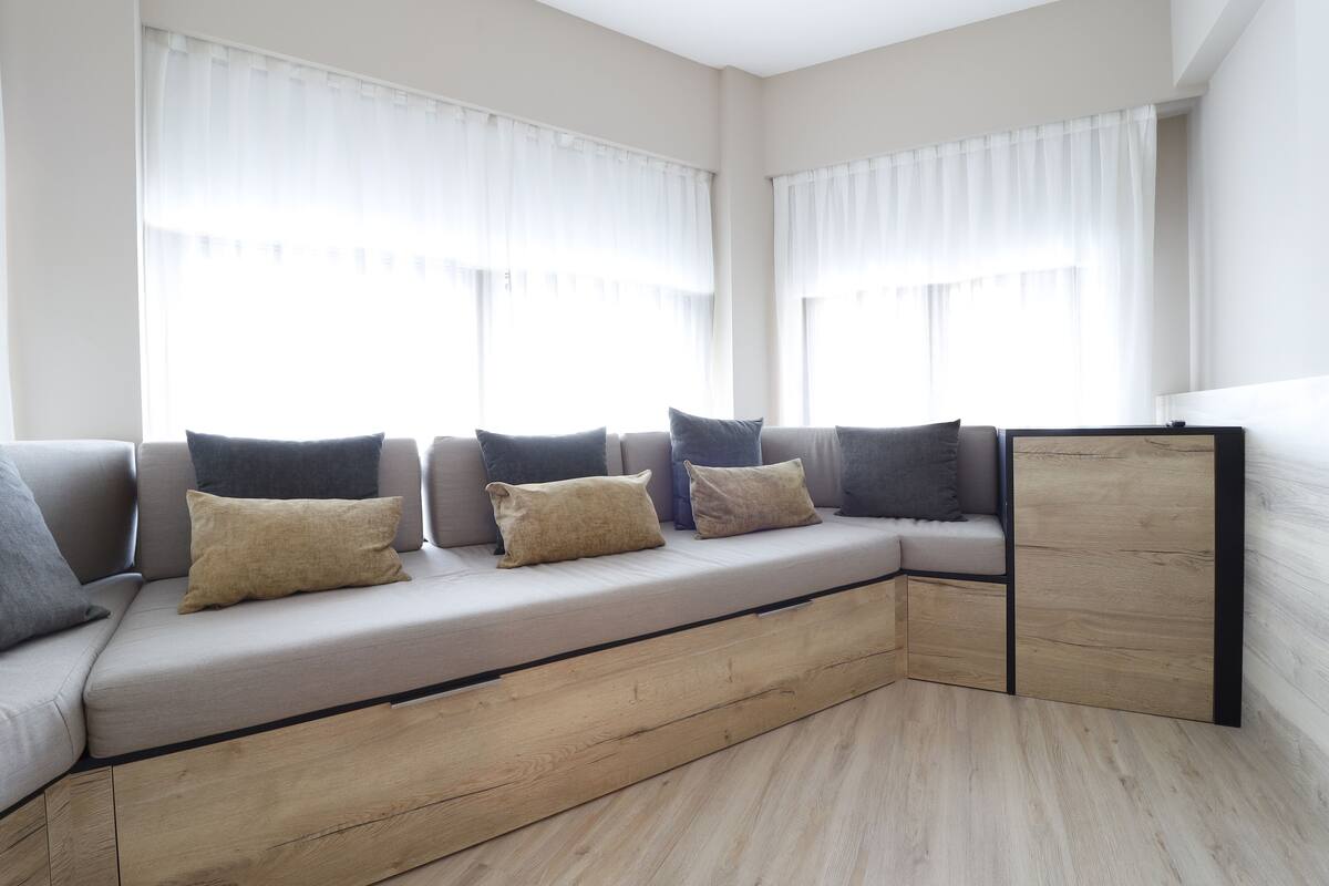 A modern living room with a built-in sofa, large windows with sheer curtains, and a wooden floor.