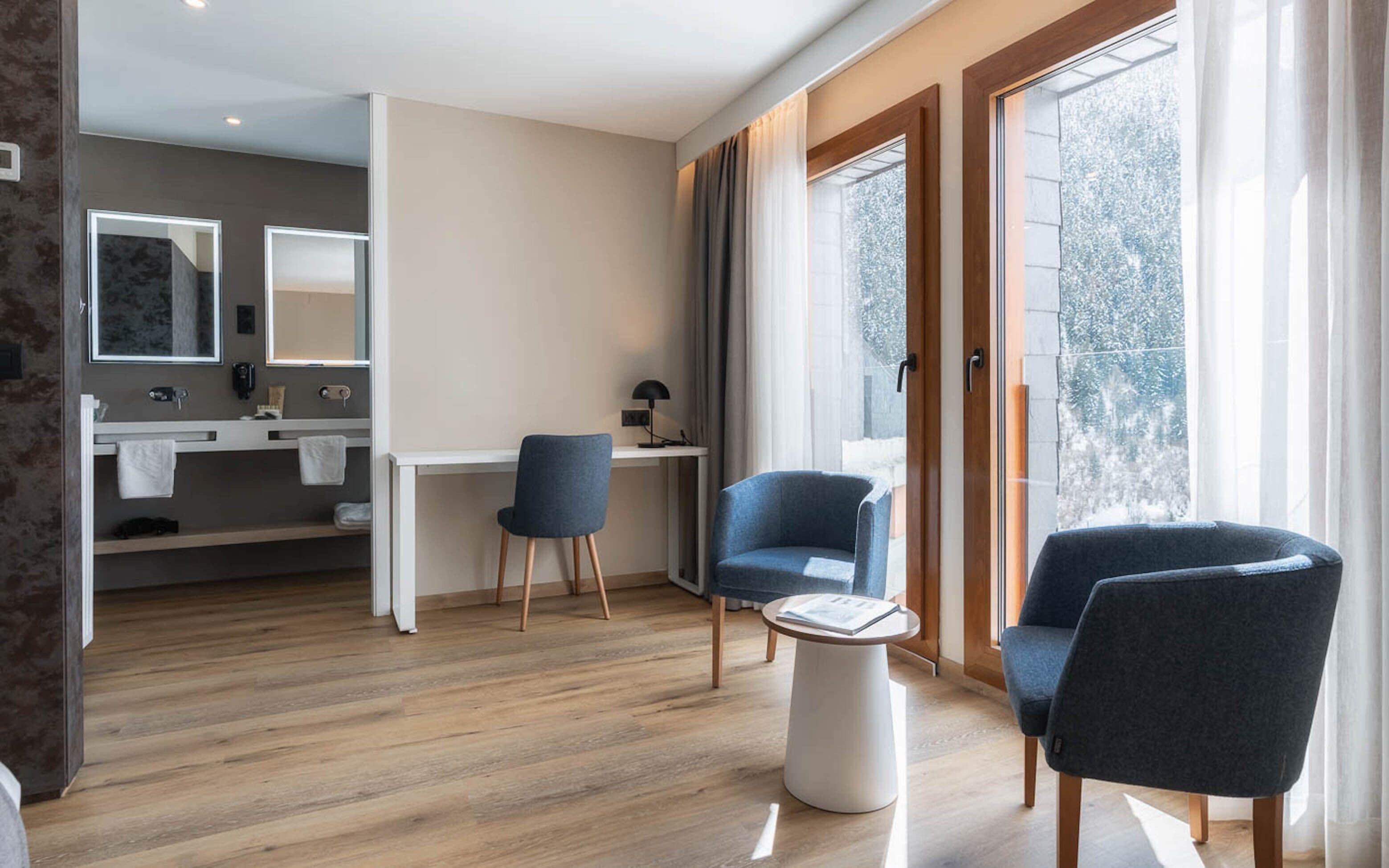 Una sala de estar moderna con piso de madera, dos sillones azules, una mesita blanca y un escritorio con una silla. Hay una ventana grande con cortinas, un espejo y un baño visible al fondo.