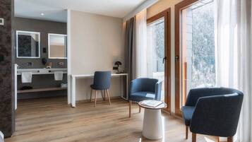 Una sala de estar moderna con piso de madera, dos sillones azules, una mesita blanca y un escritorio con una silla. Hay una ventana grande con cortinas, un espejo y un baño visible al fondo.