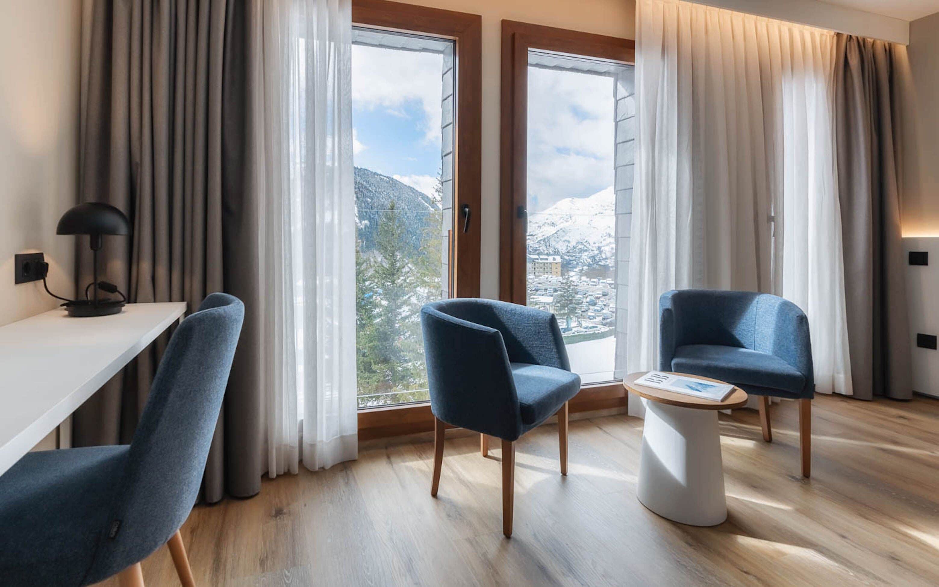 A modern hotel room with two large windows, a desk with a lamp, and two blue armchairs.