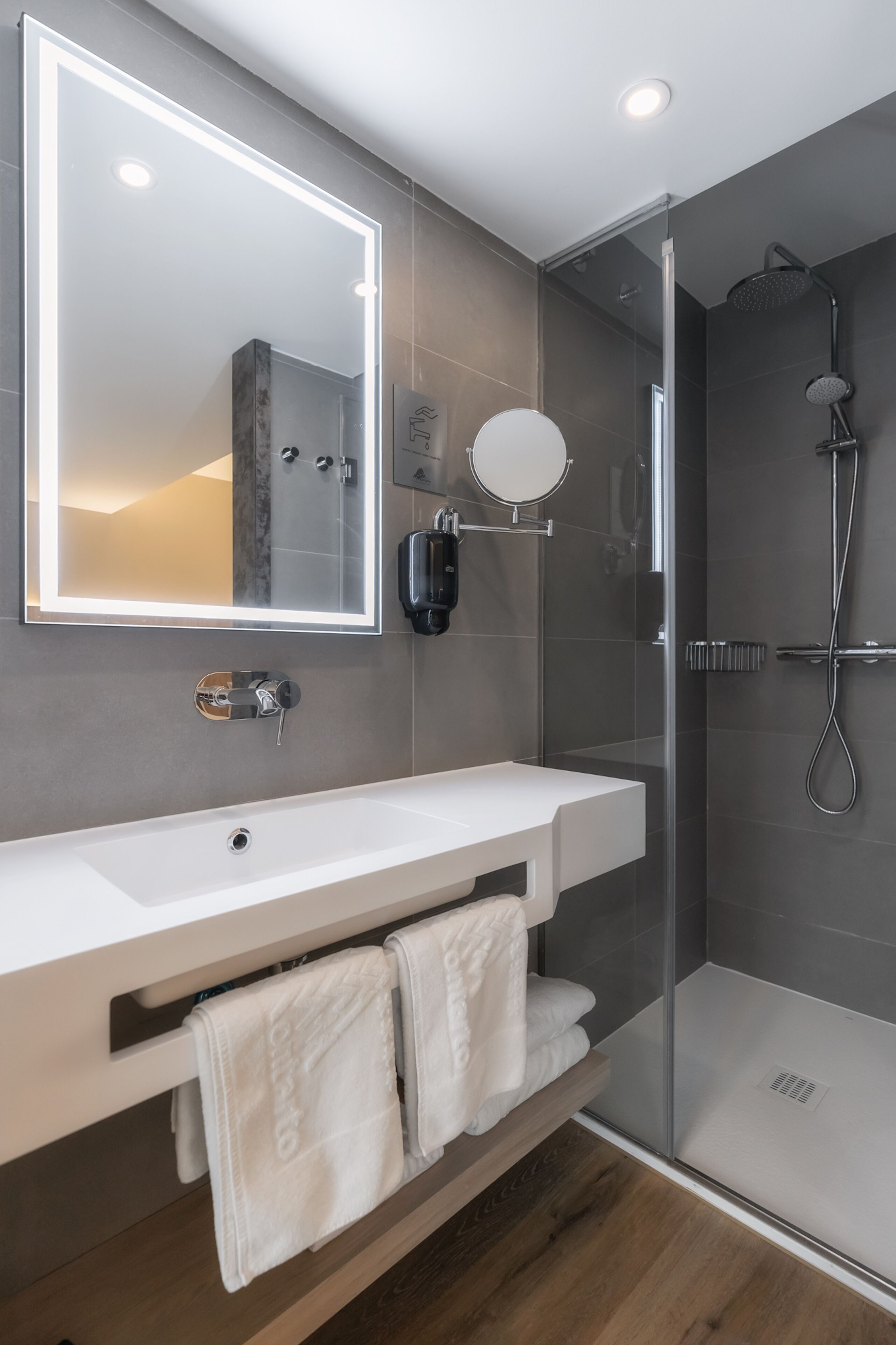 A modern bathroom with a white sink, a large mirror with LED lighting, a glass-enclosed shower, and a wooden floor.