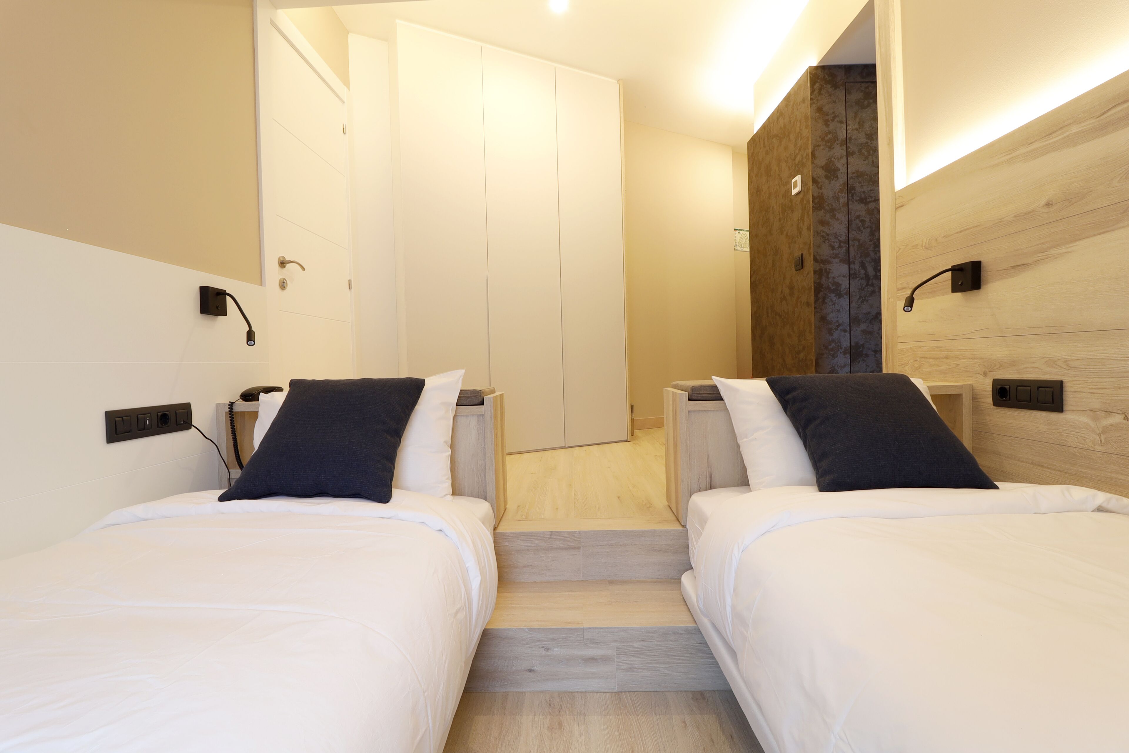 A modern hotel room with two beds, a wooden accent wall, and a small staircase leading to a door.
