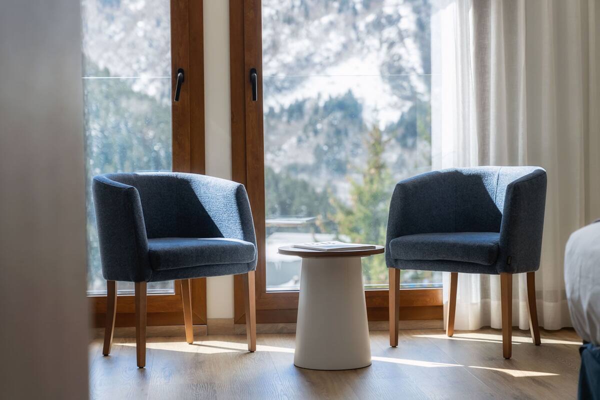 A room with two blue armchairs and a small white table, a large window with a view of mountains, and wooden flooring.