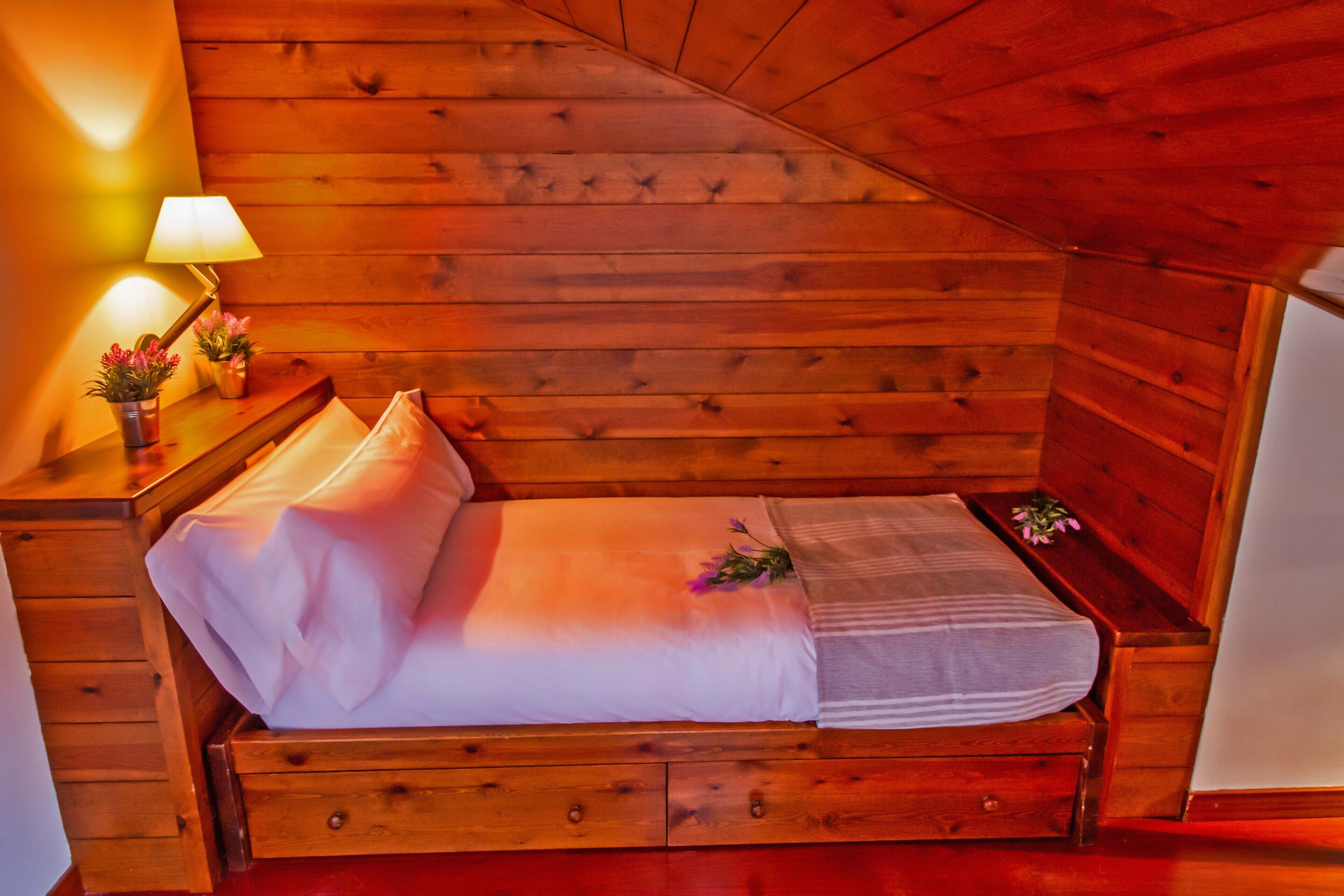 A wooden cabin-style bedroom with a single bed, a nightstand with a lamp, and a small table with flowers.