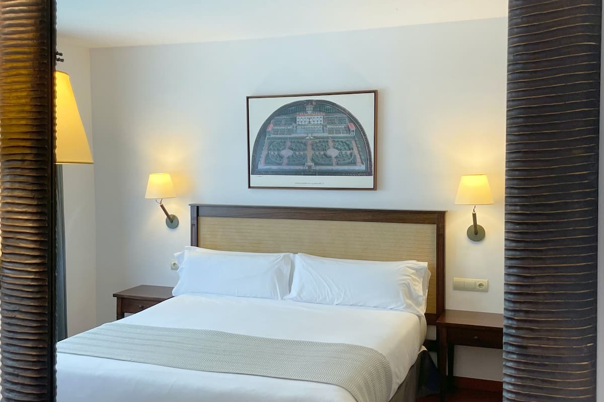 Ein Hotelzimmer mit großem Spiegel, einem Bett mit weißer Bettwäsche, zwei Nachttischlampen, einem gerahmten Bild an der Wand und einem Holz-Kopfteil.