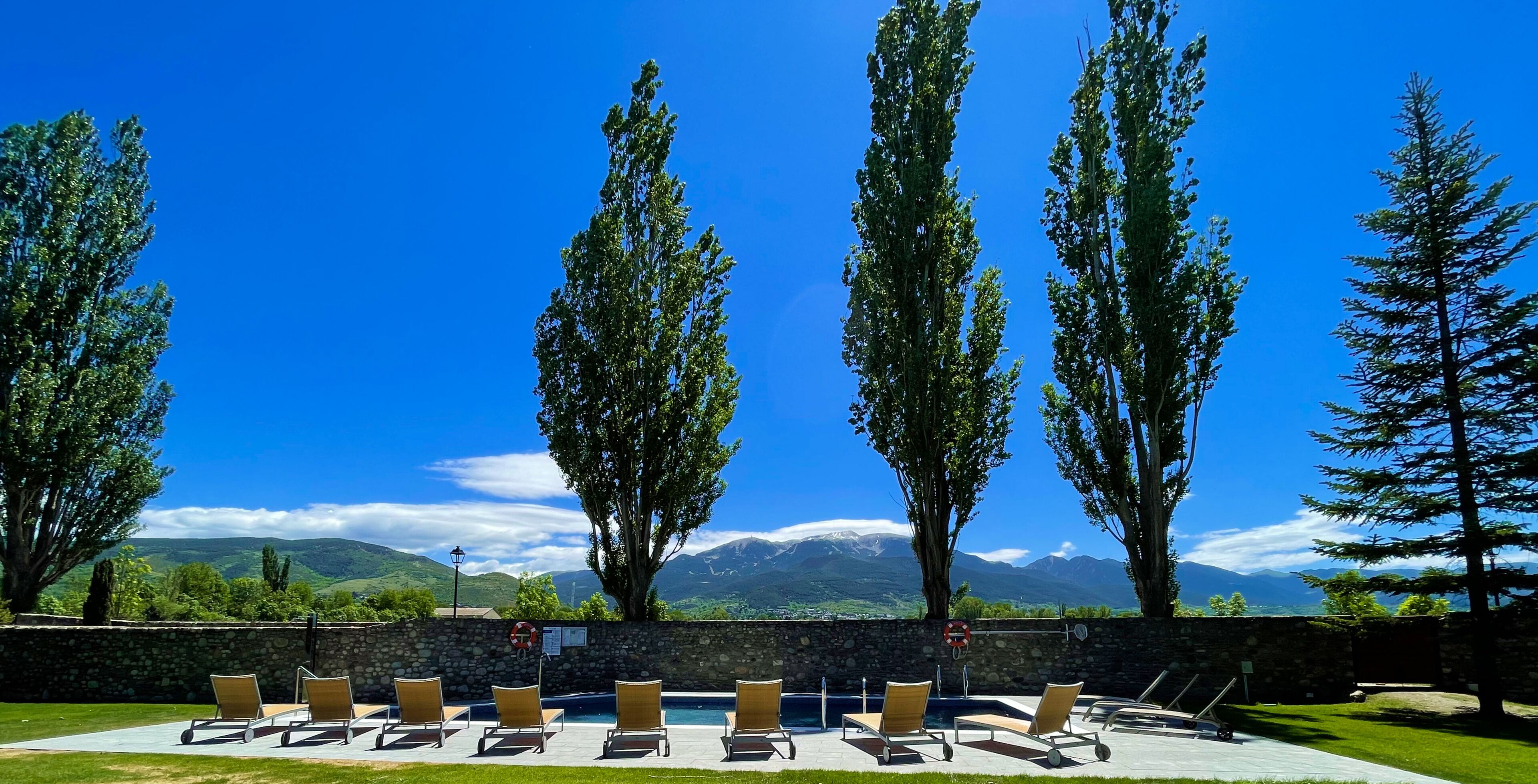 A pool area with lounge chairs and a stone wall, overlooking a mountainous landscape.