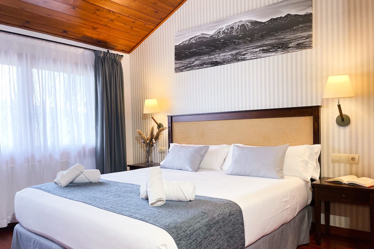 Ein Hotelzimmer mit einem großen Bett, zwei Nachttischlampen, einem Holzkopfschild, einem Fenster mit Vorhängen und einem an der Wand hängenden Bild.