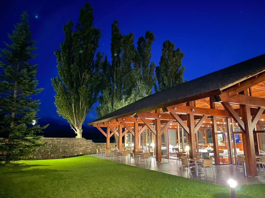 An outdoor dining area with wooden pergolas and tables, surrounded by a stone wall and trees.