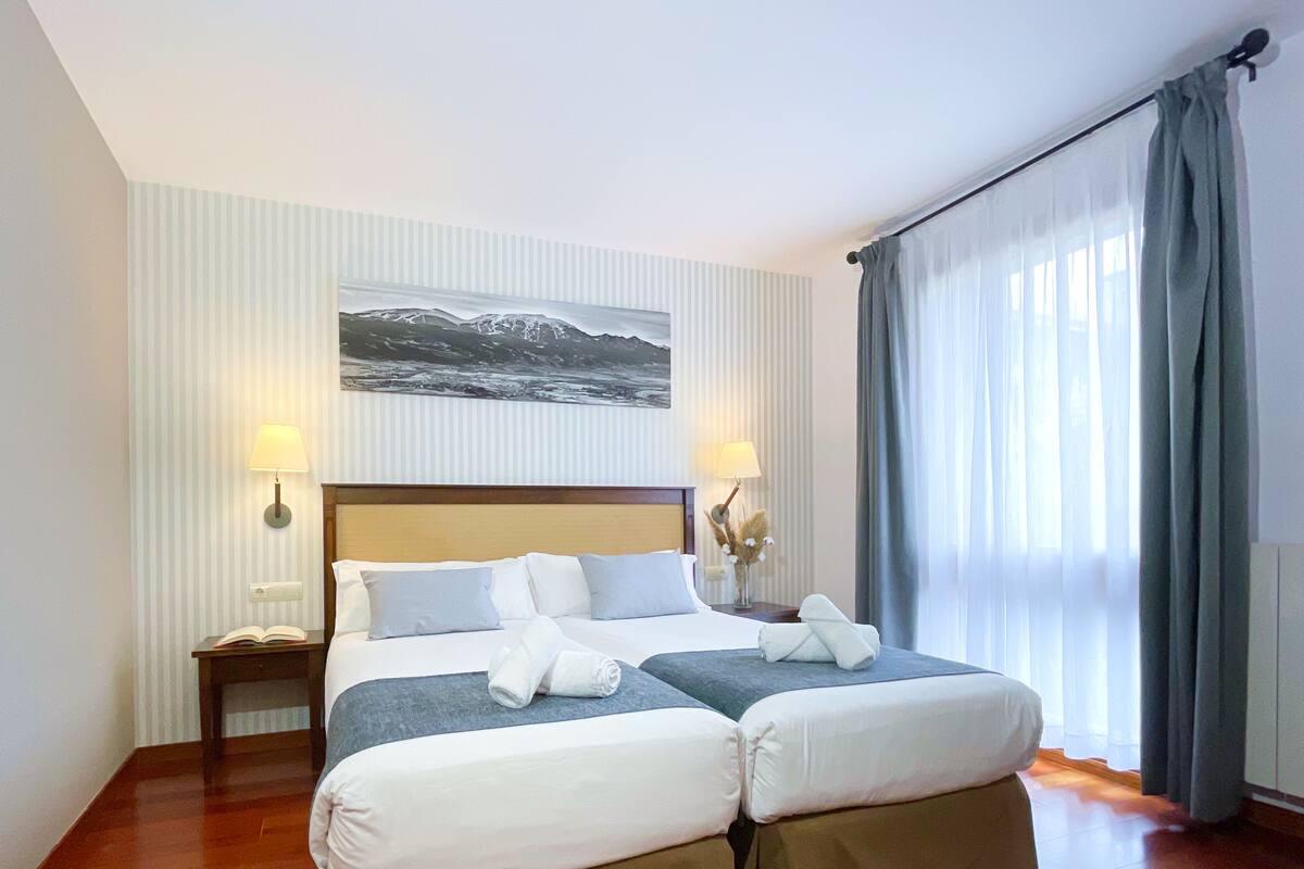 Ein Hotelzimmer mit zwei Betten, einem großen Gemälde an der Wand, einem Nachttisch mit Lampe und einem Fenster mit Vorhängen.