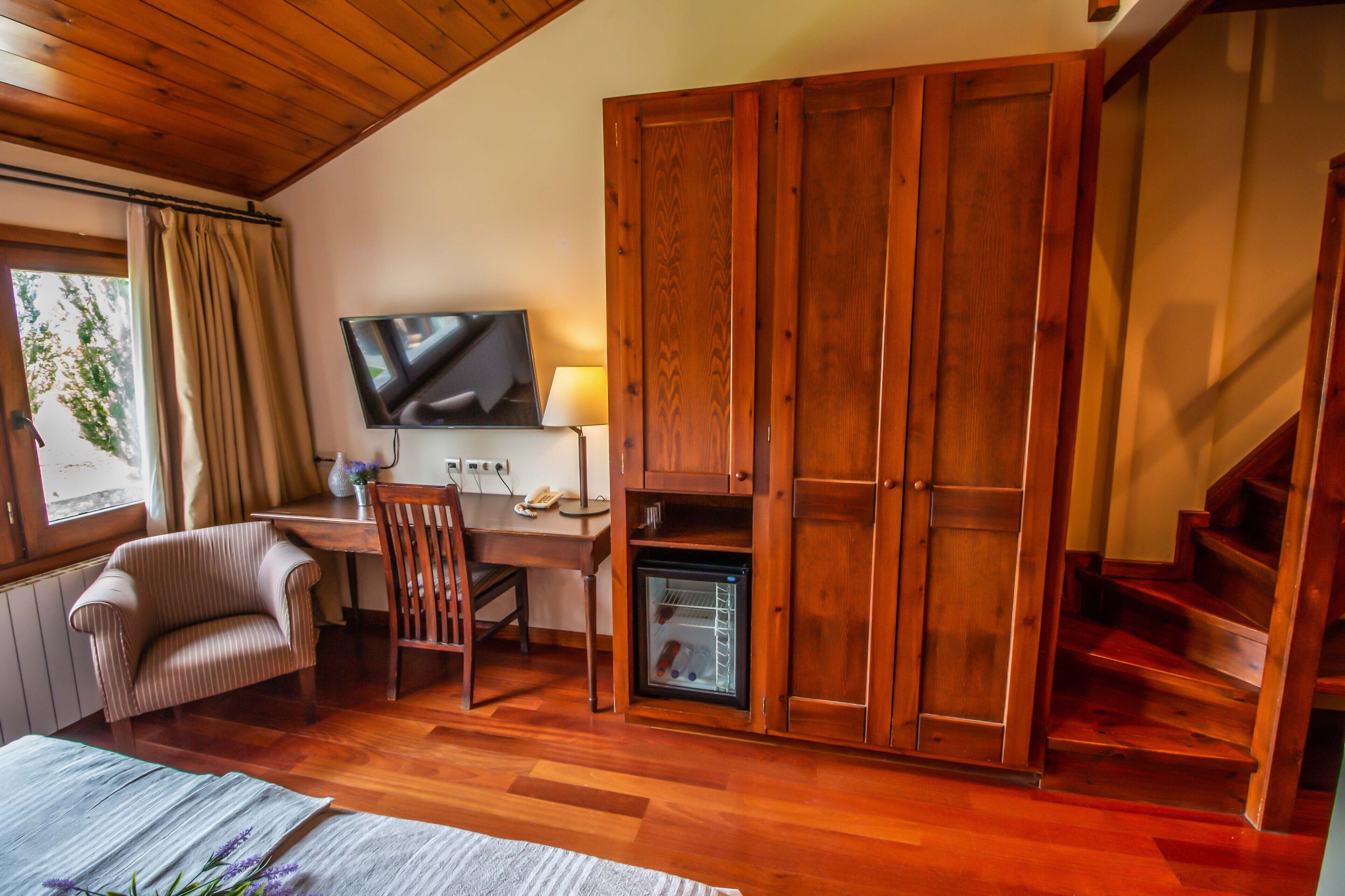 A cozy room with a wooden ceiling, a TV, a desk with a lamp, a chair, and a wardrobe.