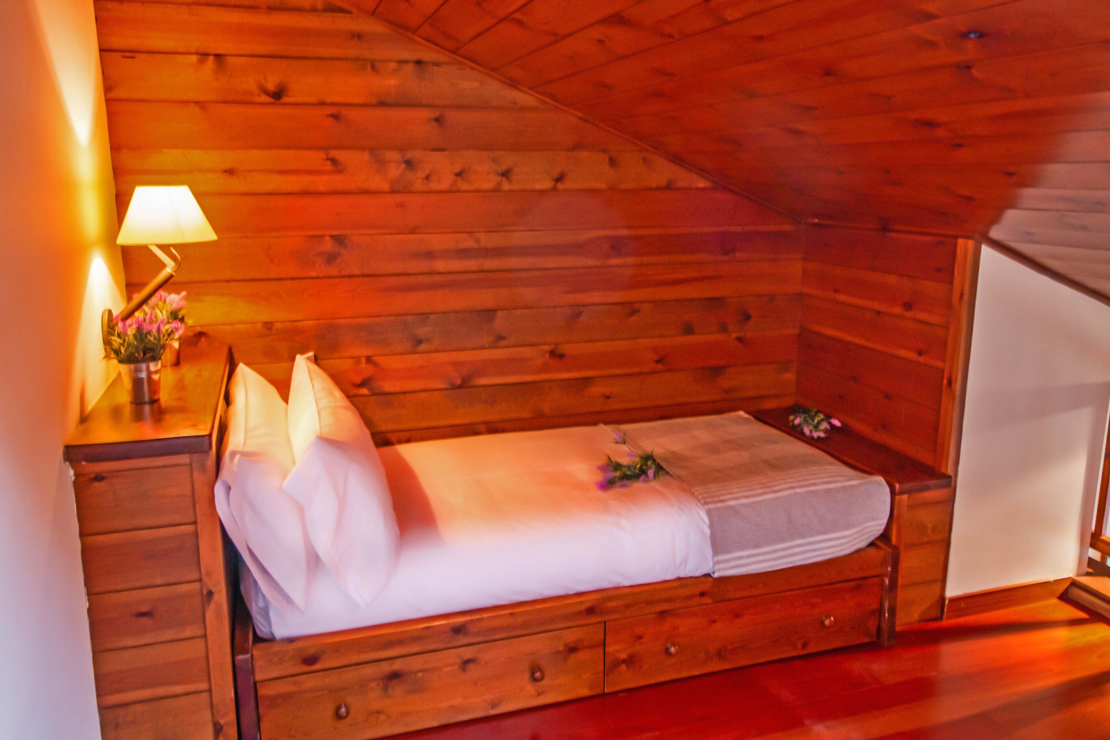 A small, cozy wooden room with a single bed, a nightstand with a lamp, and a dresser.