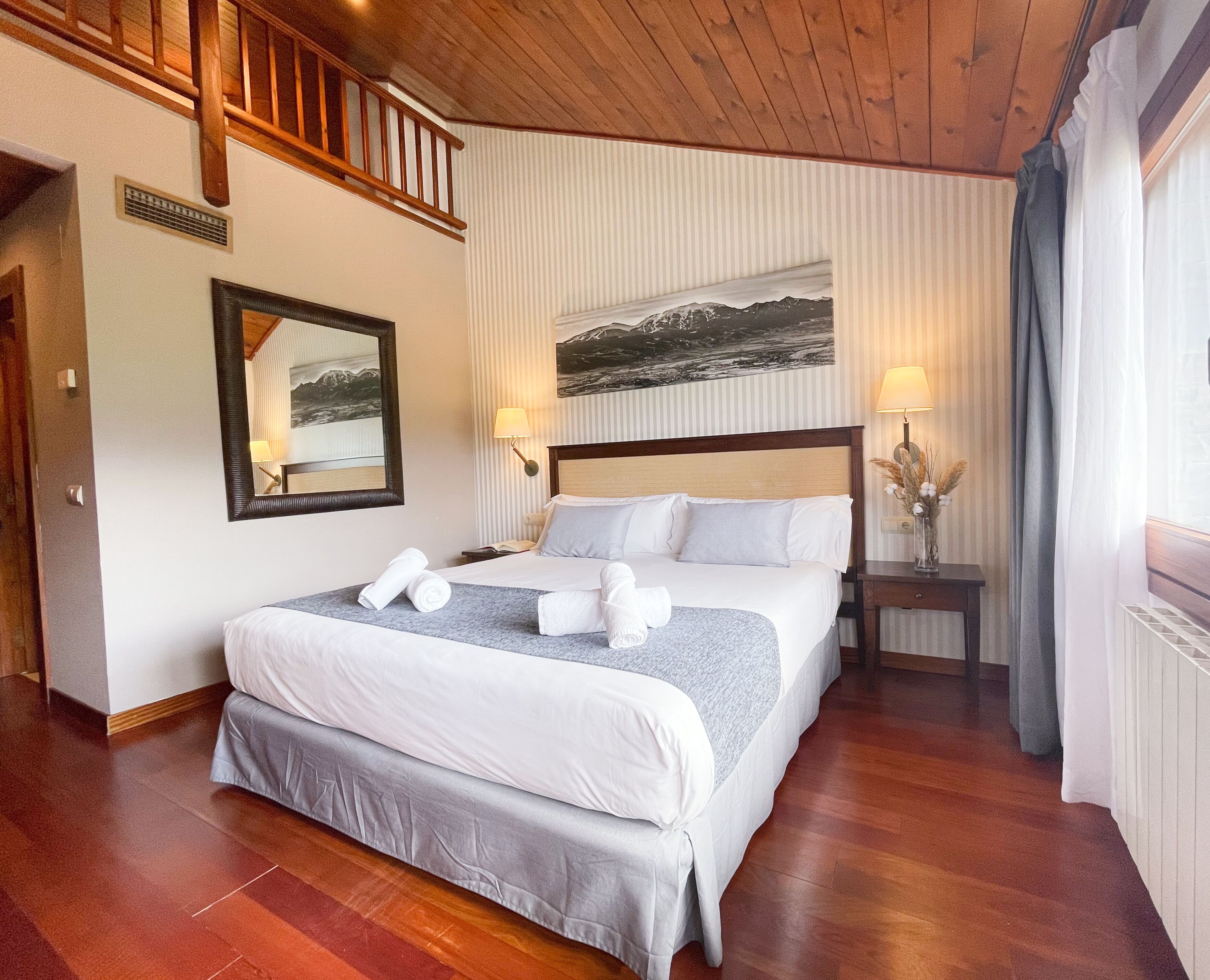 Ein Hotelzimmer mit einem großen Bett, Holzboden, einem Bild an der Wand, einem Nachttisch mit Lampe und einem Fenster mit Vorhängen.