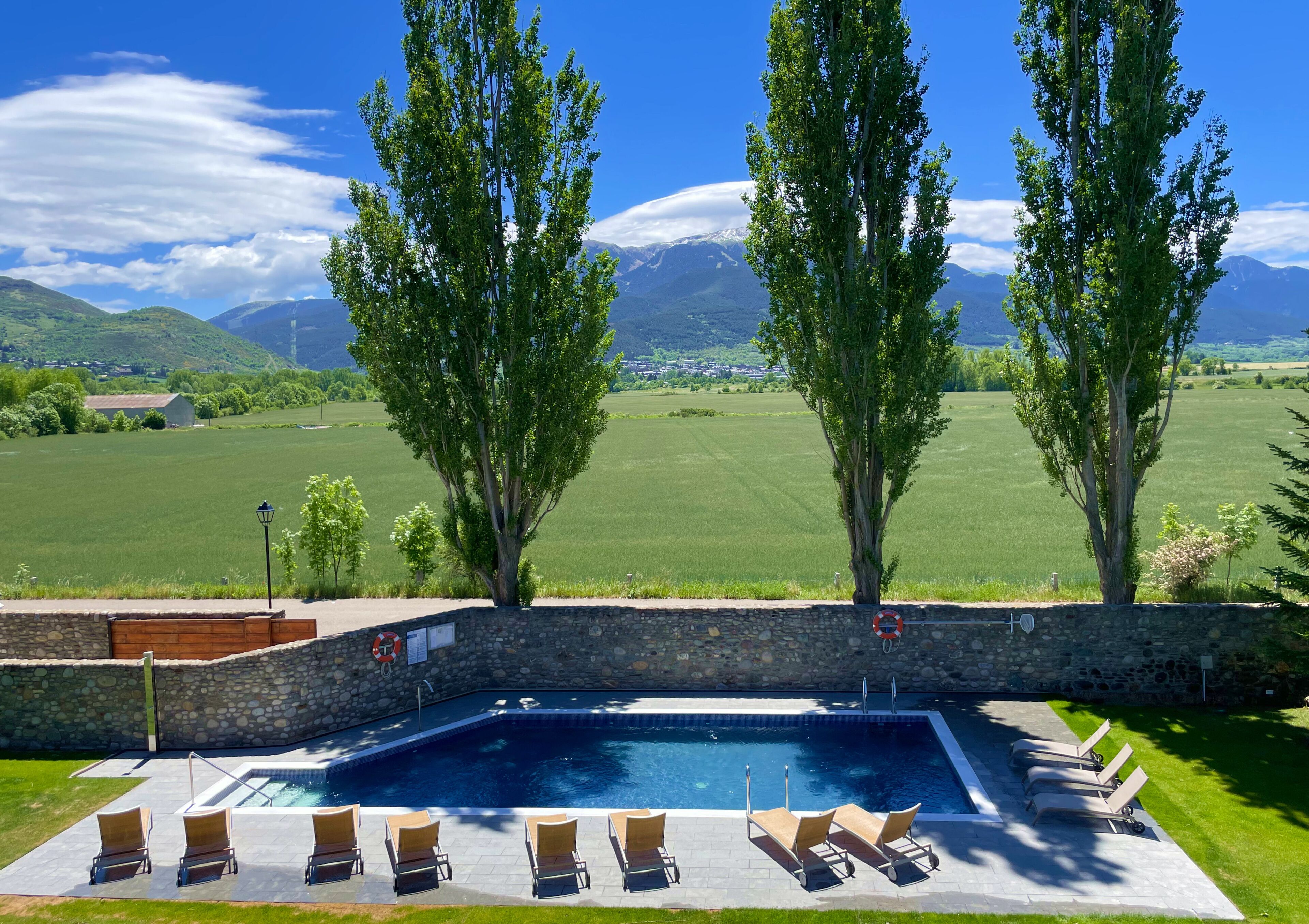 A swimming pool with lounge chairs, surrounded by a stone wall, with a view of green fields and mountains in the background.