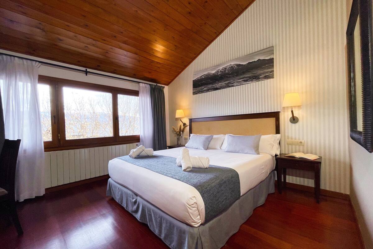 Ein Hotelzimmer mit Holzboden, einem großen Bett mit weißen Leinen, zwei Nachttischlampen, einem Fenster mit Vorhängen und einem Gemälde an der Wand.