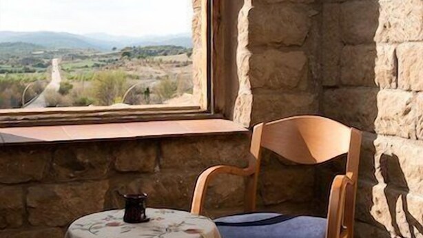 Una habitación rústica con pared de piedra, una silla de madera con cojín, una mesita con mantel estampado y vistas a un paisaje rural a través de una ventana.