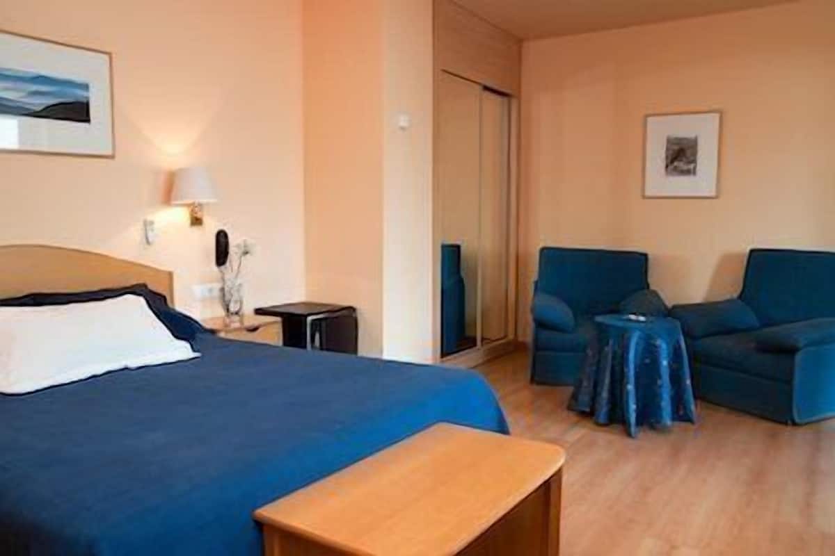 A hotel room with a bed, a desk, two armchairs, a small table, and a door.