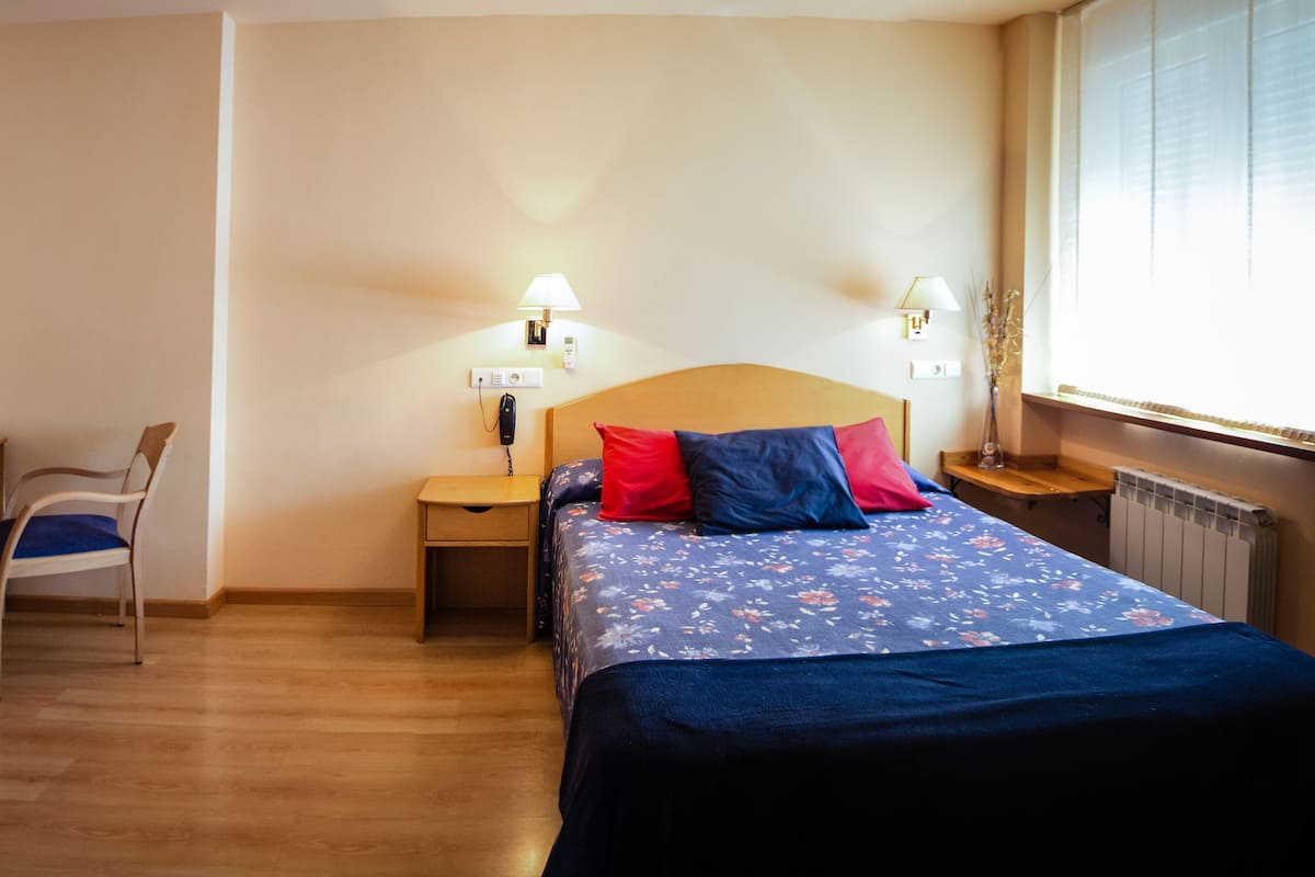 A hotel room with a bed, a desk, a chair, and a nightstand with a lamp.