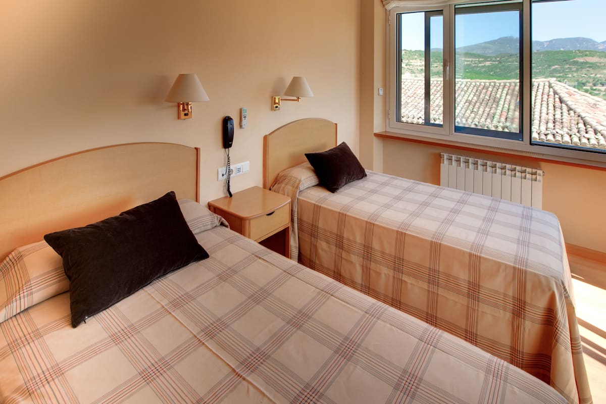 A hotel room with two beds, a nightstand, a lamp, a telephone, and a window with a view of the countryside.