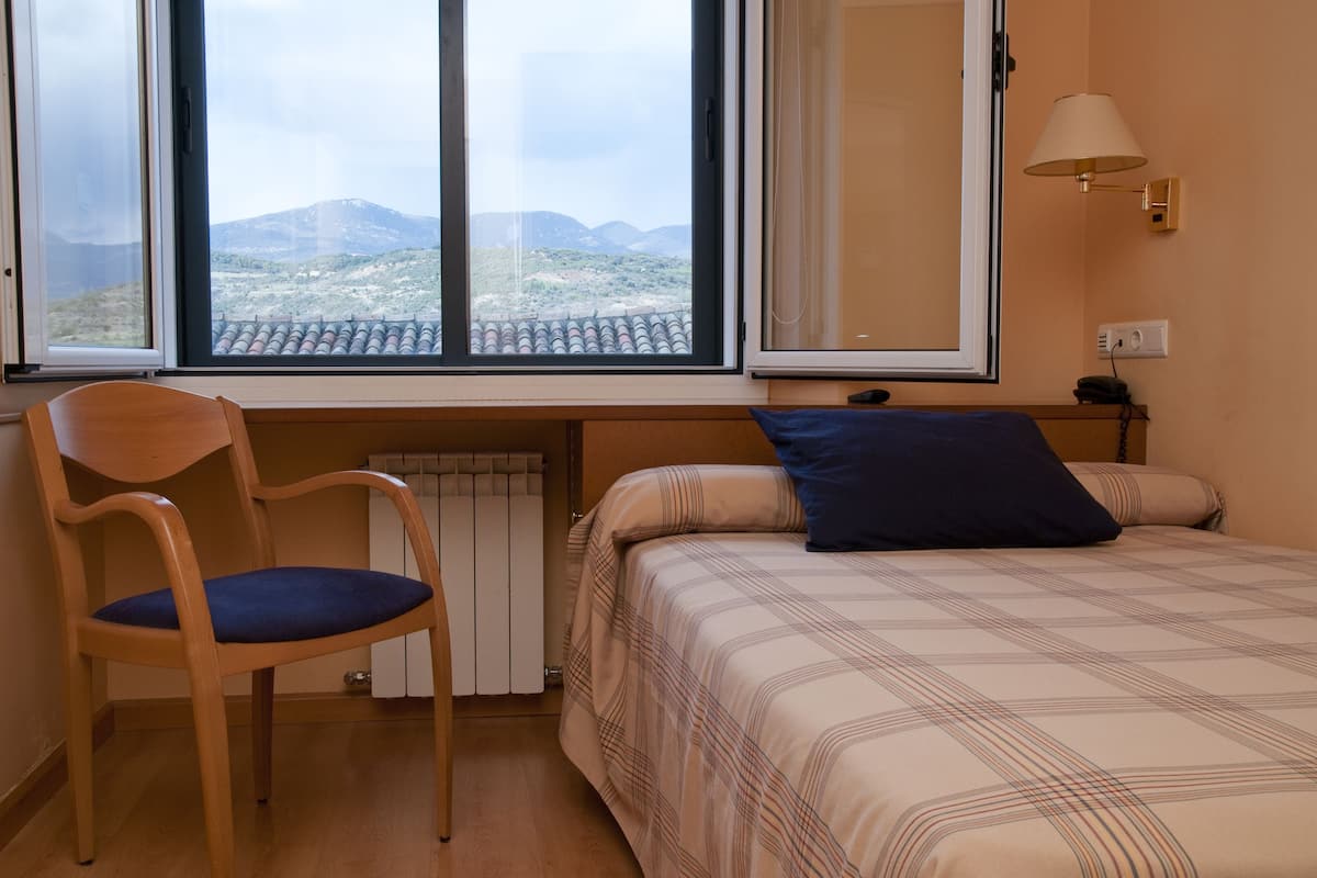A bedroom with a bed, a chair, a window with a view of mountains, and a lamp.