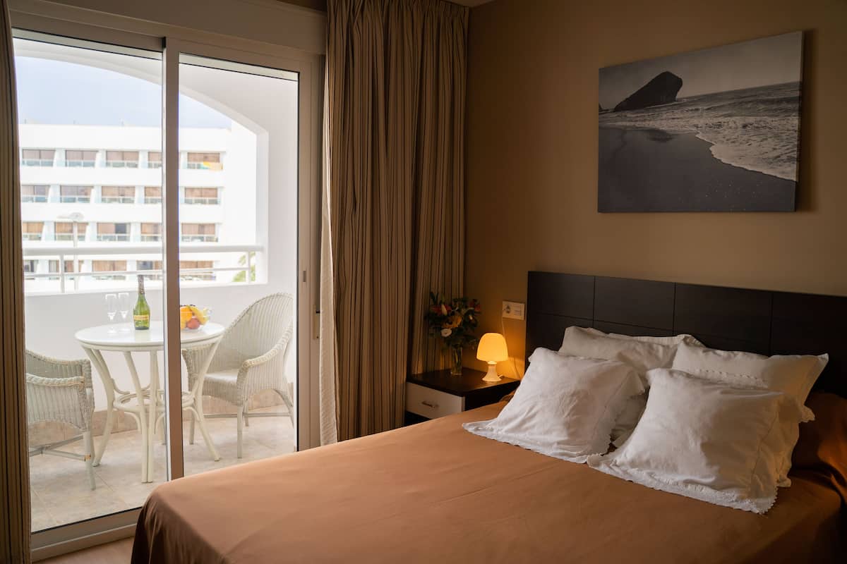 A hotel room with a bed, a nightstand with a lamp, a window with curtains, a balcony with a table and chairs, and a painting on the wall.