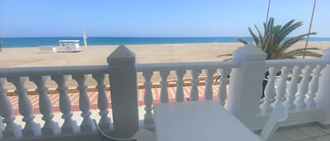 A balcony with a view of the beach, a table, and chairs.