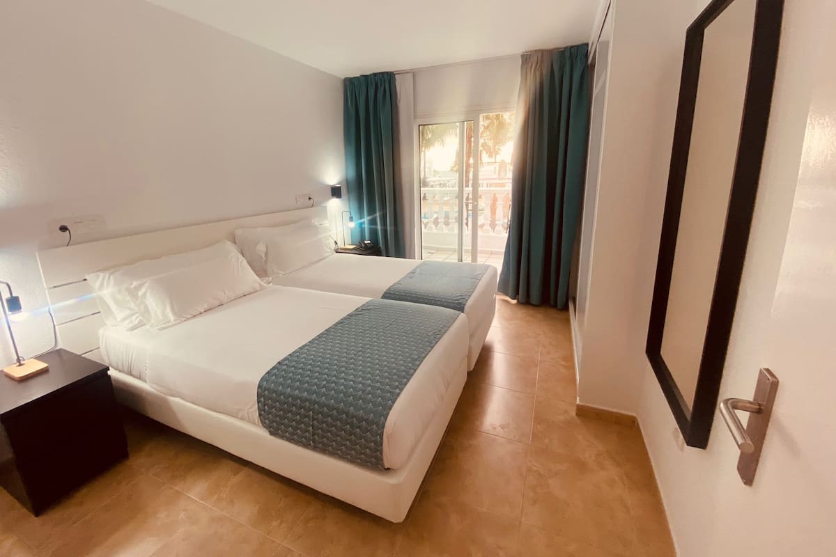 A hotel room with two beds, a nightstand, a lamp, and a view of the outdoors through a window with curtains.