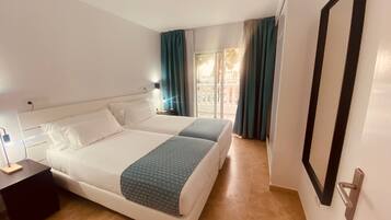 Quarto de hotel com duas camas, uma mesa de cabeceira, uma luminária e vista para o exterior através de uma janela com cortinas.