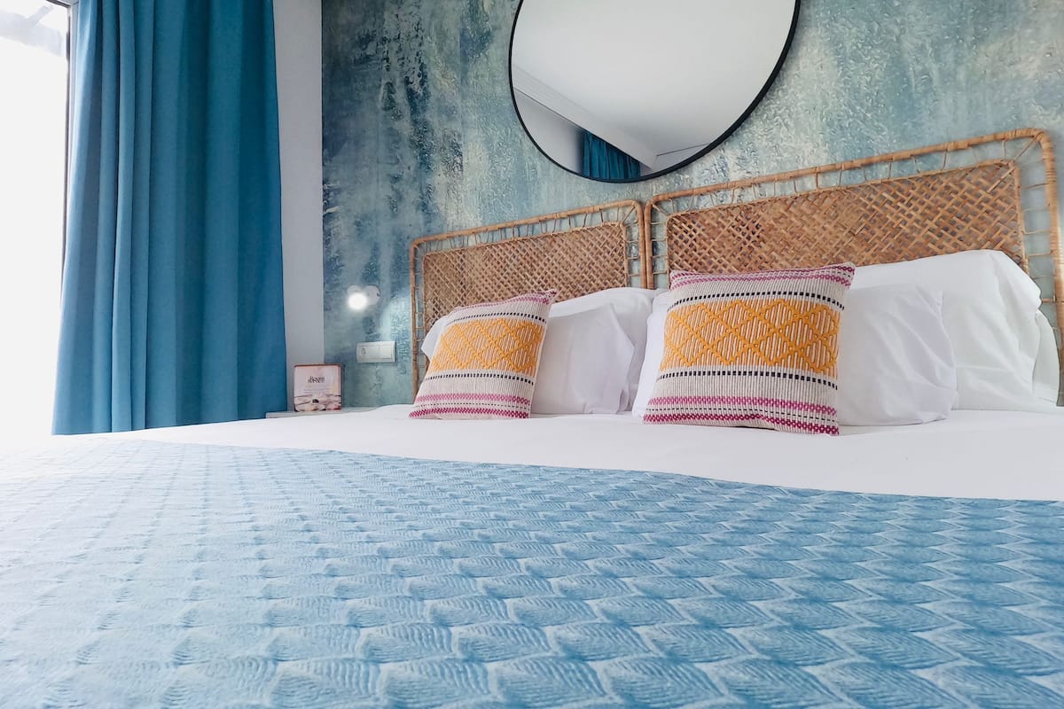 A bed with a blue patterned bedspread, a round mirror above it, and a textured wall behind.