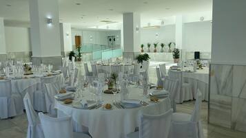 Un salón de banquetes con mesas redondas dispuestas para cenar, manteles blancos y sillas con fundas blancas.