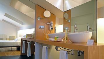 Un baño moderno con un espejo grande, un lavabo y un secador de pelo. En la habitación contigua hay una cama.