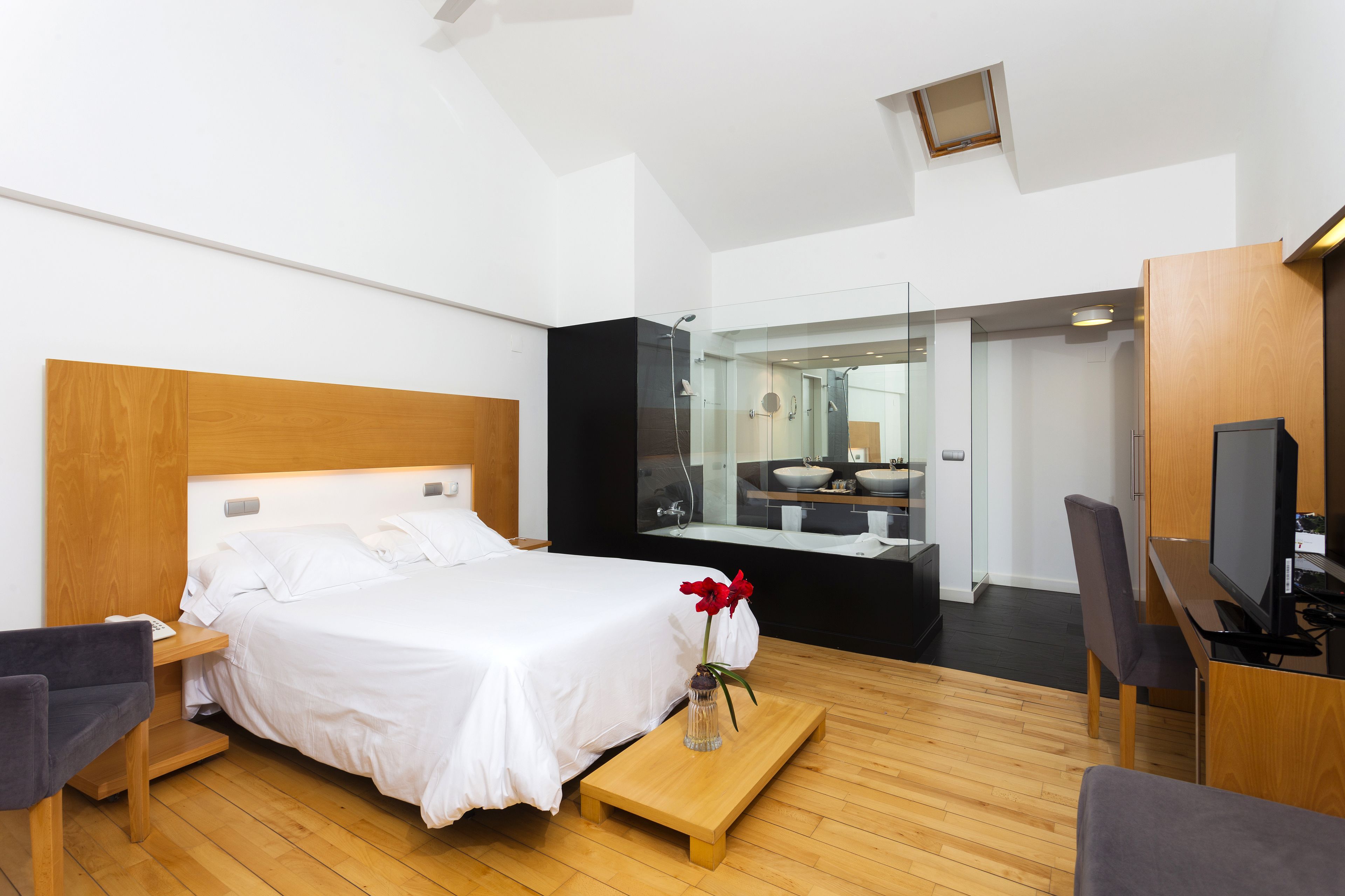 Una camera d'albergo moderna con un letto grande, un comodino in legno, un tavolino con un vaso, una TV e un bagno visibile tramite una porta in vetro.