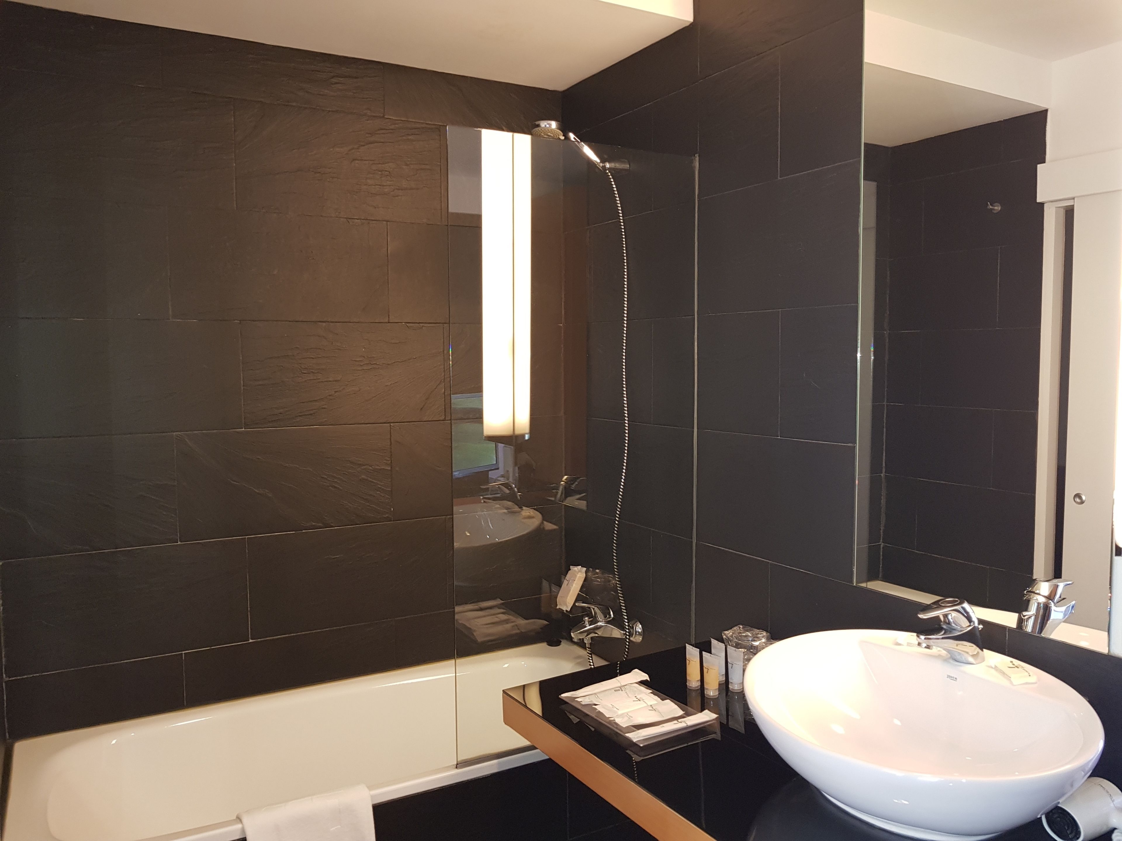 A modern bathroom with dark tiled walls, a white bathtub, a large mirror, and a sink with toiletries.