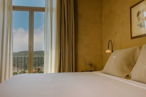 Una camera d'albergo con un letto, un comodino, un quadro incorniciato, una finestra con tende e vista su un paesaggio montano.