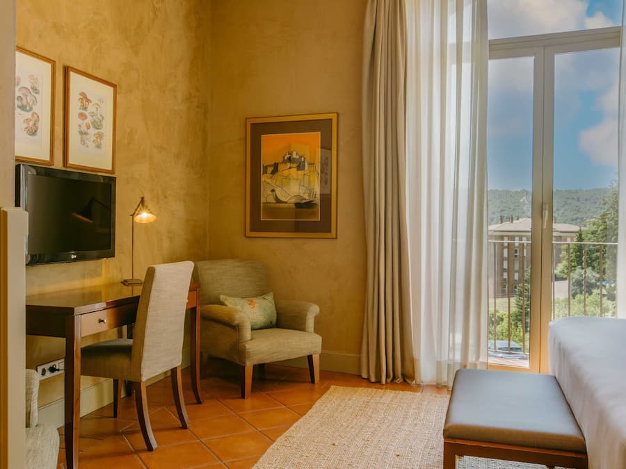 Quarto de hotel com escrivaninha, cadeira, televisão, cama e vista para o exterior.