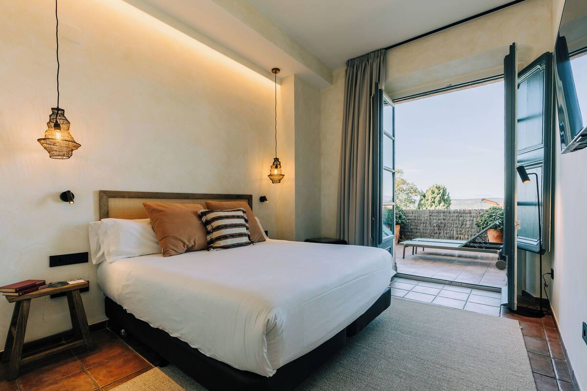 Une chambre moderne avec un grand lit, deux lampes de chevet, une petite table avec un livre, et une vue sur l’extérieur grâce à une porte coulissante.