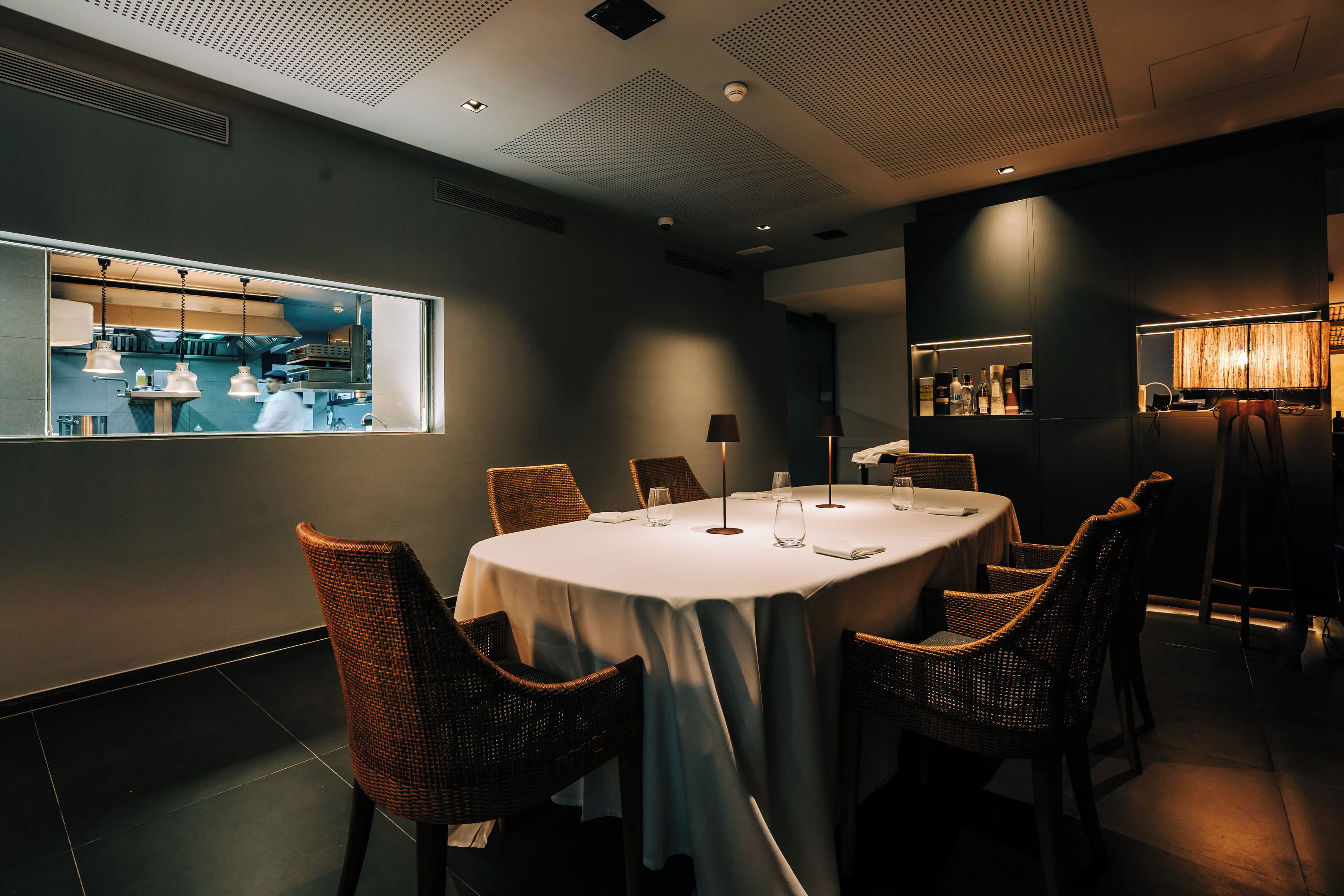 A modern dining room with a round table, wicker chairs, and a view of the kitchen through a large window.