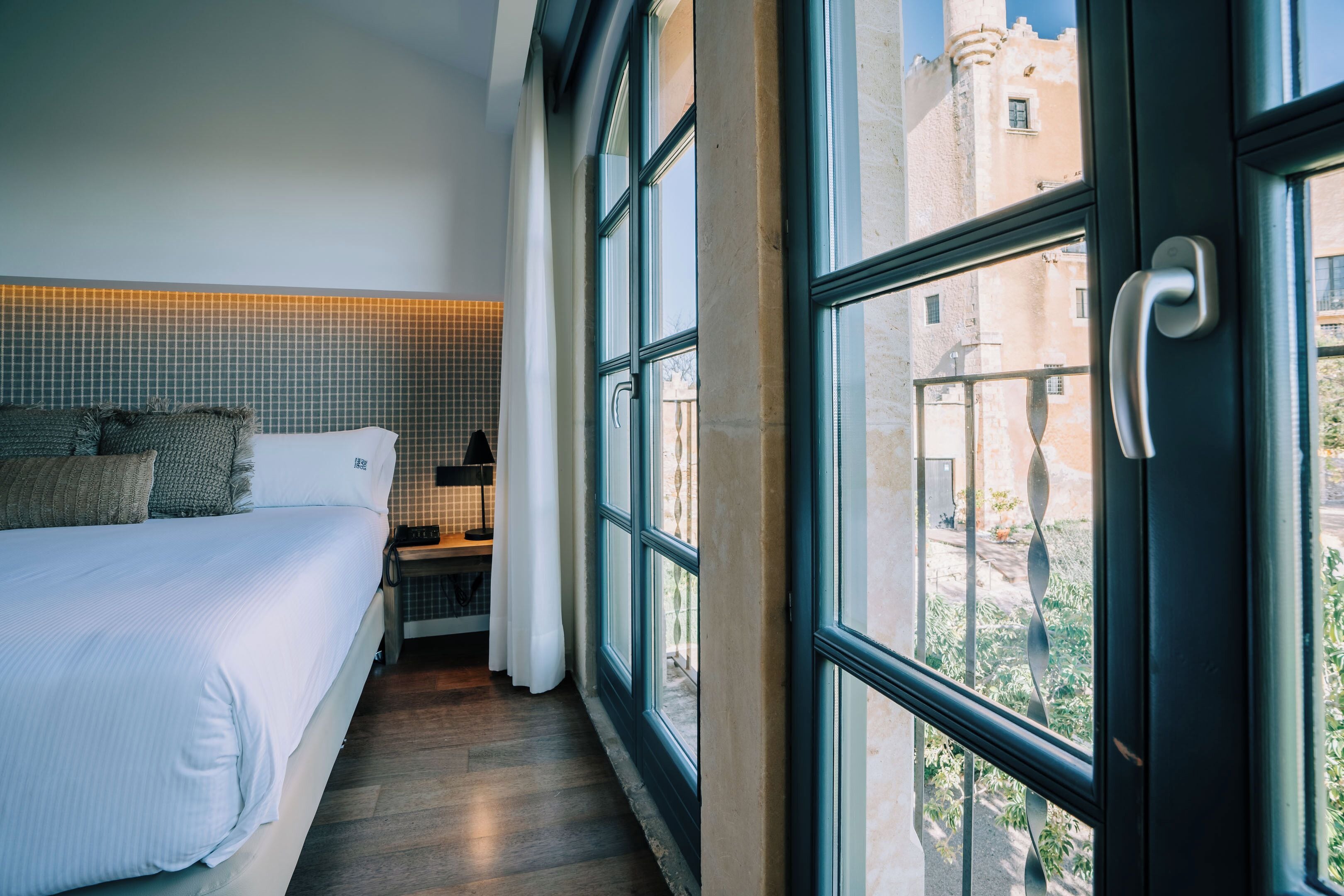 Ein modernes Hotelzimmer mit einem großen Bett, einem Nachttisch, einer Lampe und Blick auf die Stadt durch große Fenster.