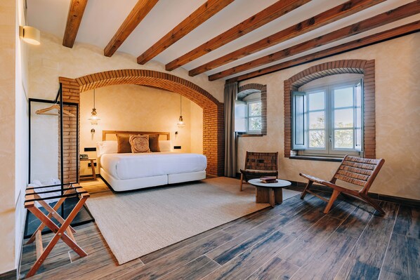 Un dormitorio con una cama grande, una silla de madera, una mesita y una ventana.