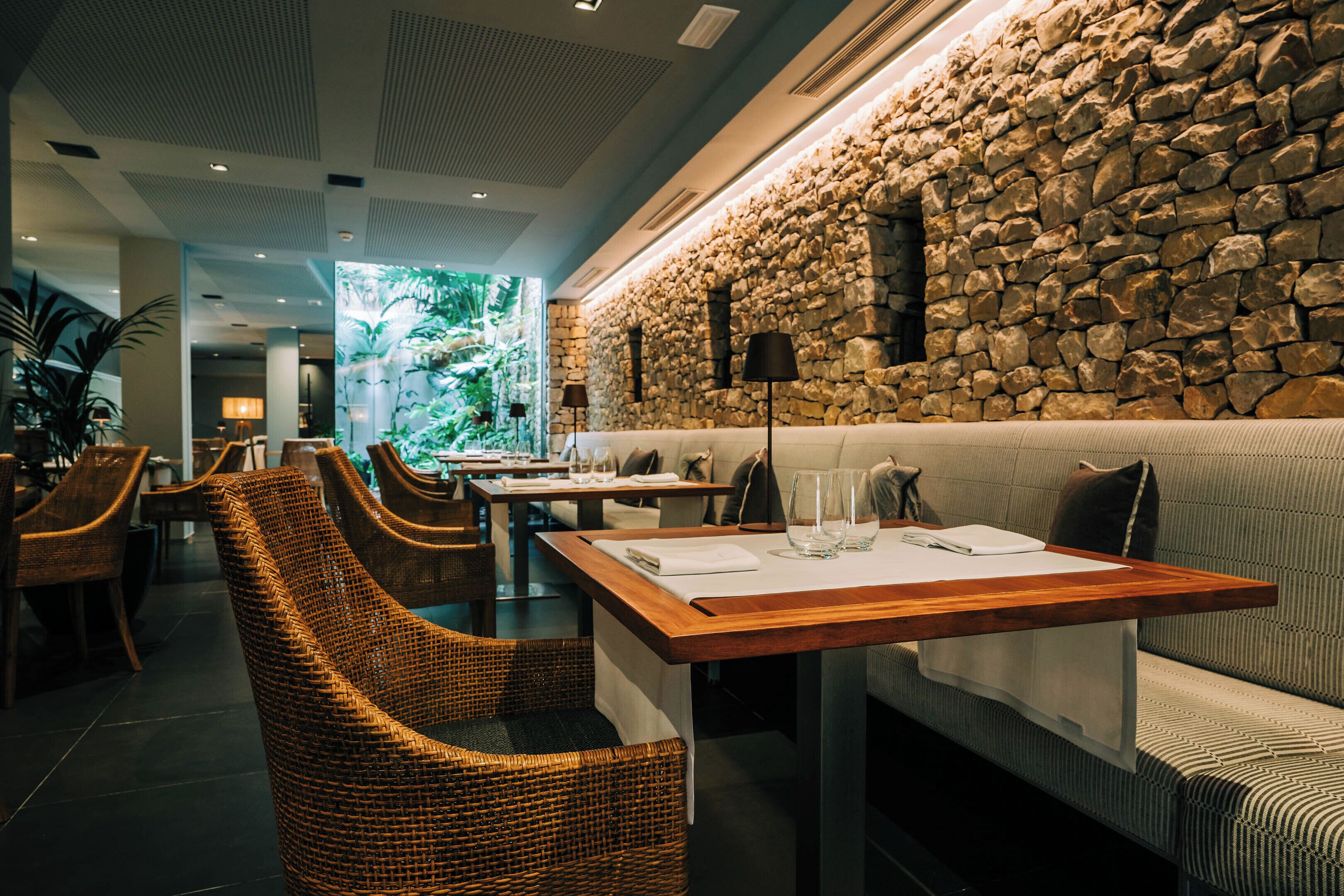 A restaurant with wicker chairs, a stone wall, and a wooden table with a white tablecloth.